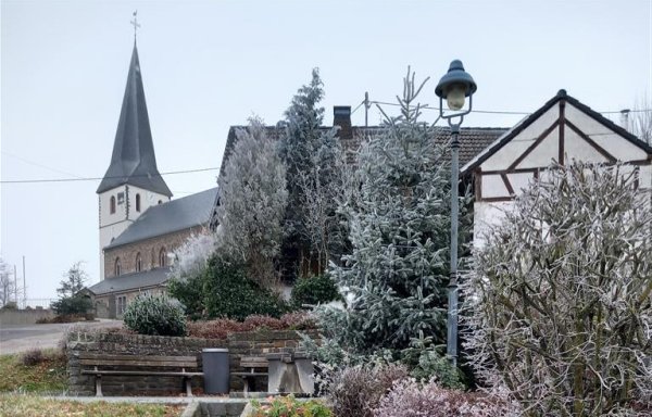 Ein kleiner Steinweg führt zum Dorfplatz mit Sitzbank, neben der ein Weihnachtsbaum platziert ist. Im Hintergrund ist die Kirche mit ihrem markanten Kirchturm zu sehen. Die gesamte Szene vermittelt eine ruhige, winterliche Atmosphäre.